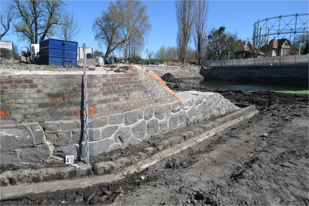 Odsłonięte murowane fundamenty XIX-wiecznej Bramy Stoczniowej podczas badań archeologicznych w Gdańsku. Widoczna tyczka geodezyjna i kamienno-ceglana konstrukcja.